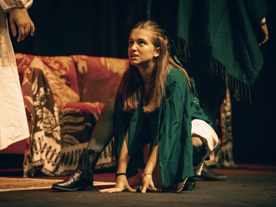 Manon Maindivide on stage in a green cloak, crouching during a theatre performance