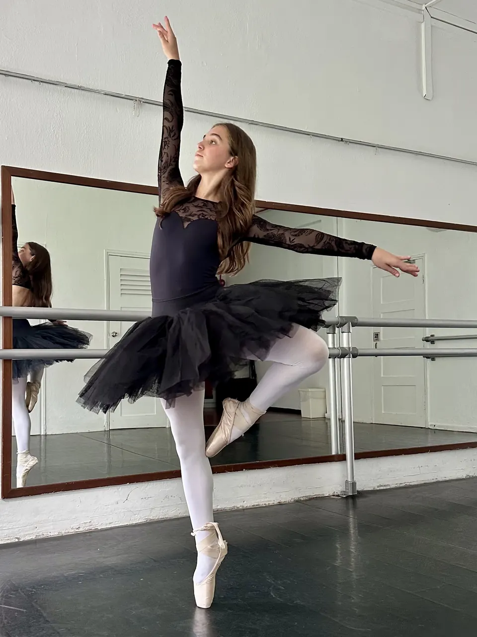 Manon Maindivide dance rehearsal en pointe in a black tutu, one arm raised high near the barre