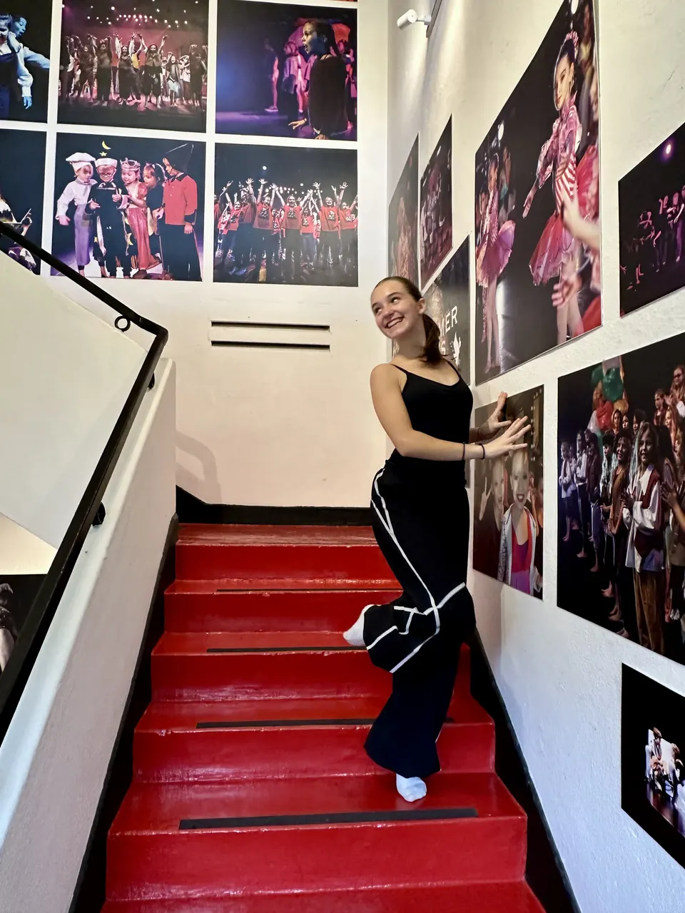 Manon Maindivide behind the scenes on a red staircase lined with show photos, dressed in black