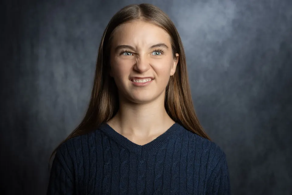 Manon Maindivide headshot in a navy cable-knit sweater, playful grin against a grey backdrop