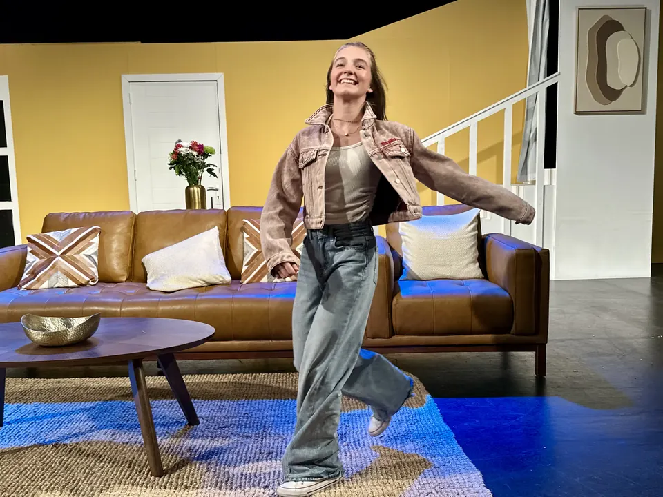Manon Maindivide on stage in a denim jacket, dancing across a living room set with a brown sofa
