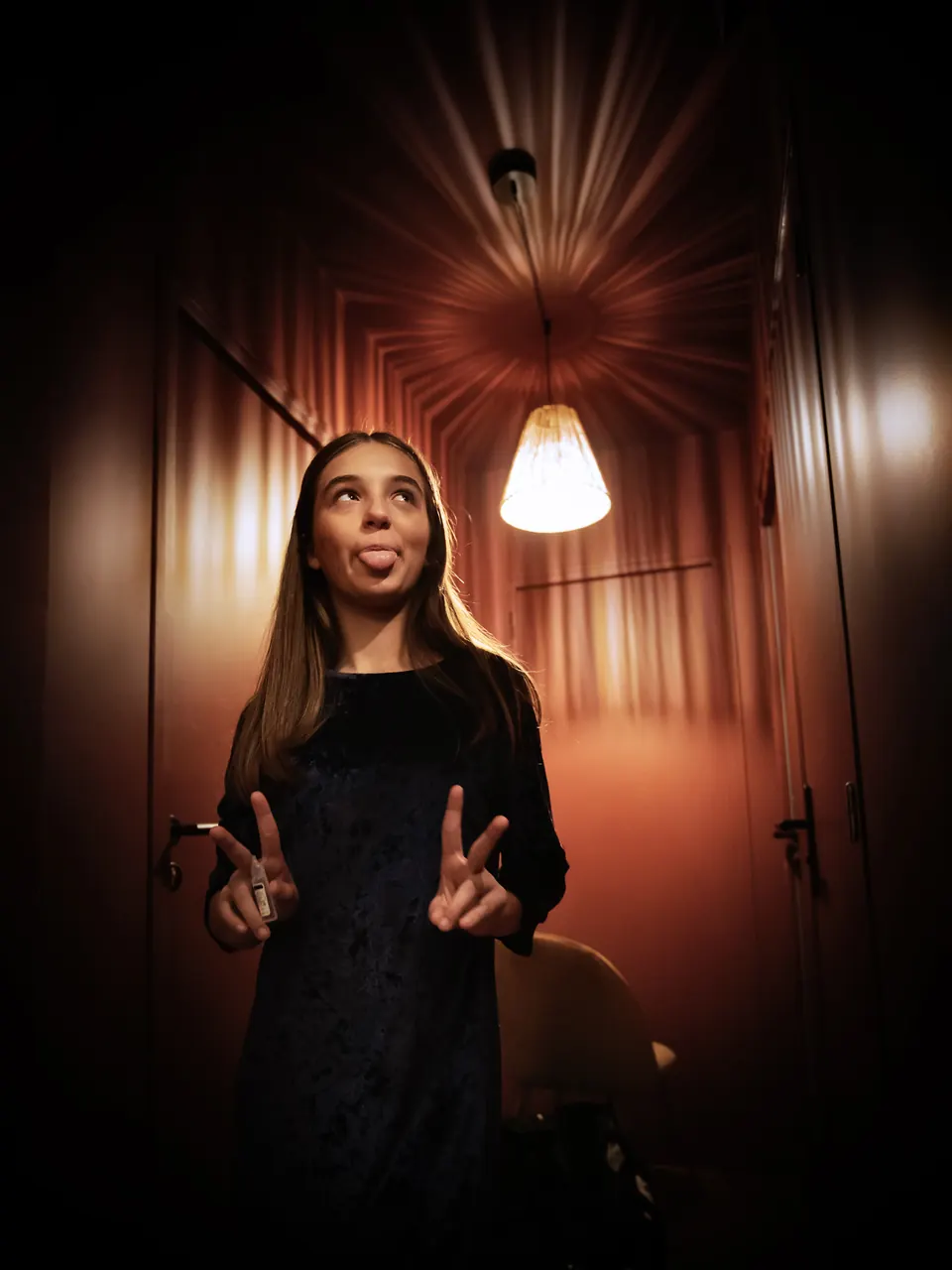 Manon Maindivide casual photo in a black dress making peace signs in a warmly lit corridor