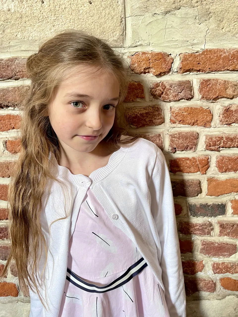 Manon Maindivide casual portrait leaning on a red brick wall, wearing a white cardigan and pink skirt