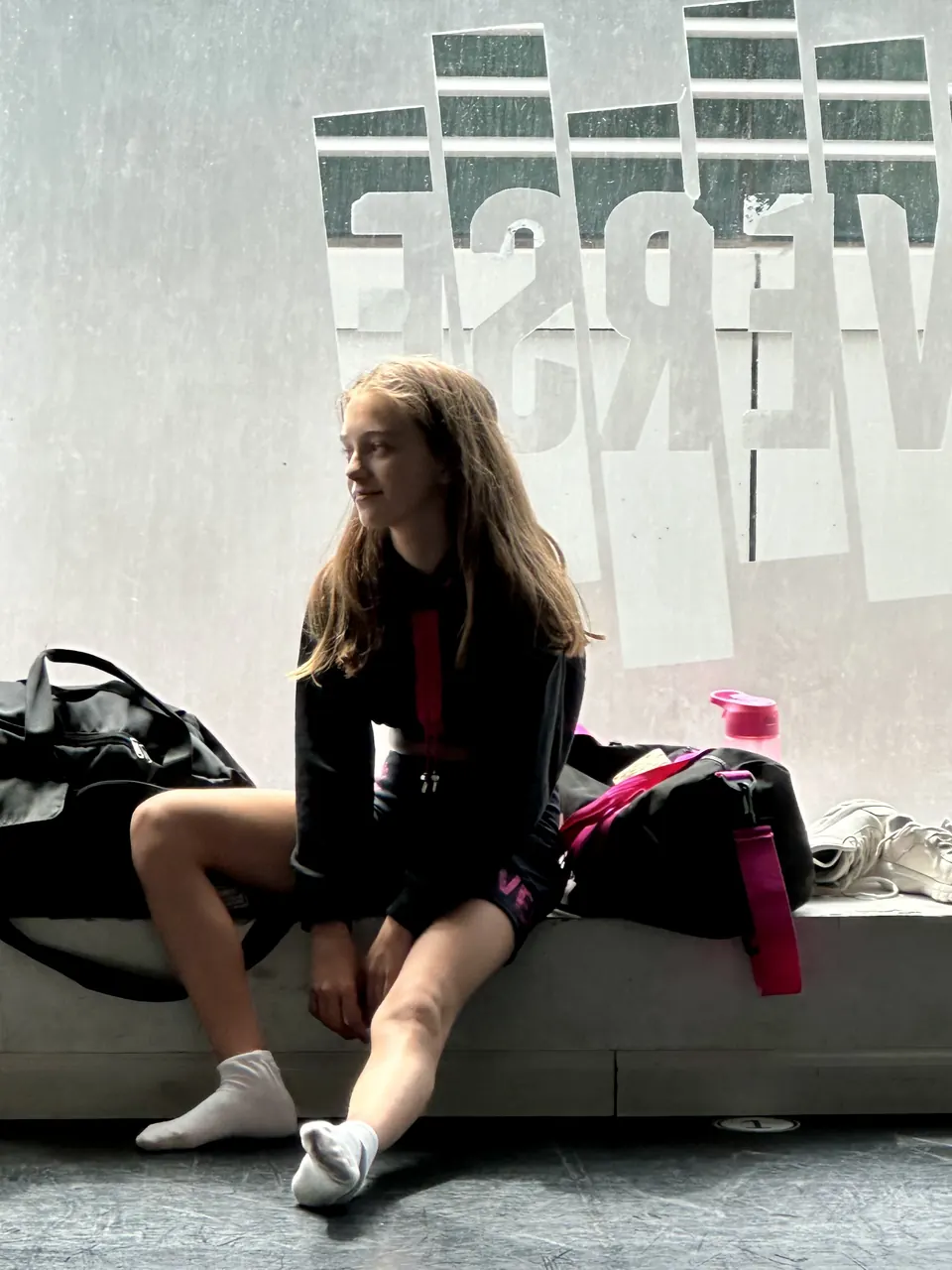 Manon Maindivide behind the scenes, sitting on a bench in a dance studio lobby with her bags
