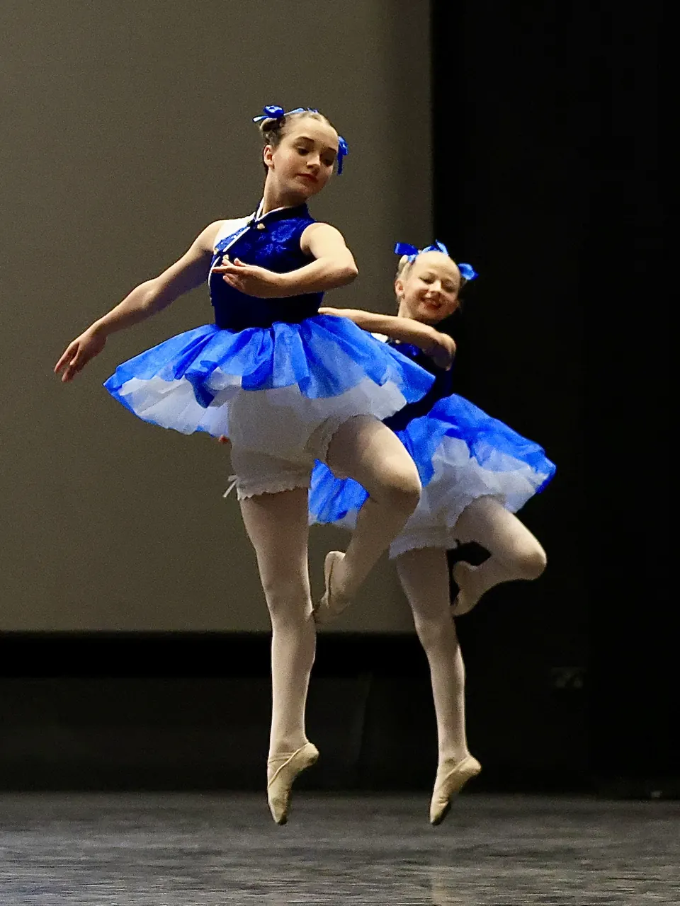 Manon Maindivide dance performance in a blue tutu with a hair bow, leaping beside another dancer
