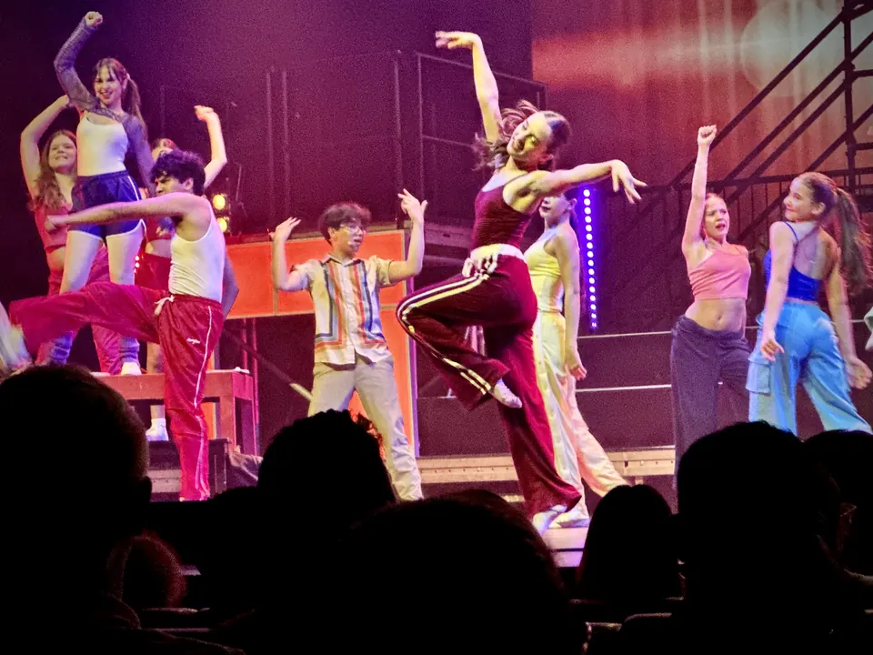 Manon Maindivide dance performance with a colourful ensemble on a lit stage, arms raised