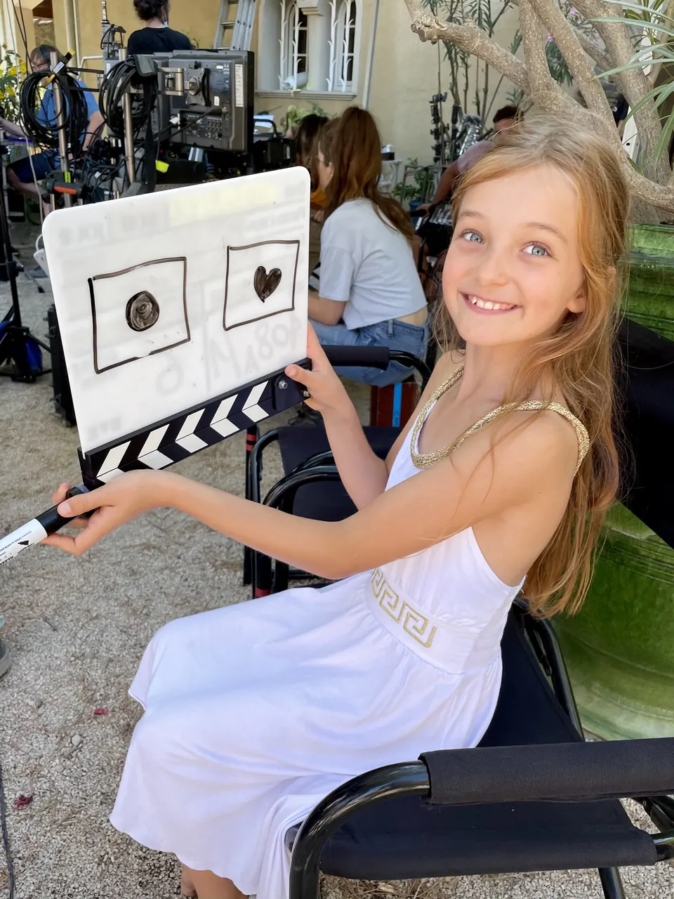 Manon Maindivide behind the scenes in a white dress, smiling and holding a clapperboard