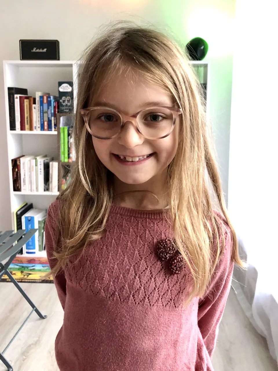 Manon Maindivide casual photo in round glasses and a pink knit sweater, smiling near a bookshelf
