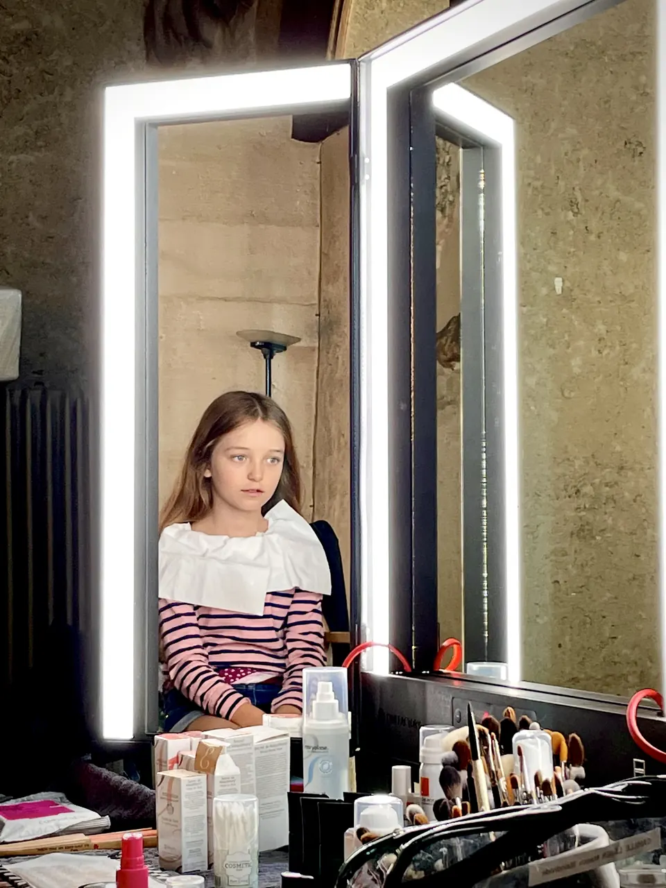 Manon Maindivide behind the scenes in a striped top, seated at a lit makeup mirror