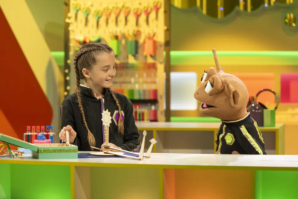 Manon Maindivide on set of a children's TV show, braided hair, at a table beside a puppet character