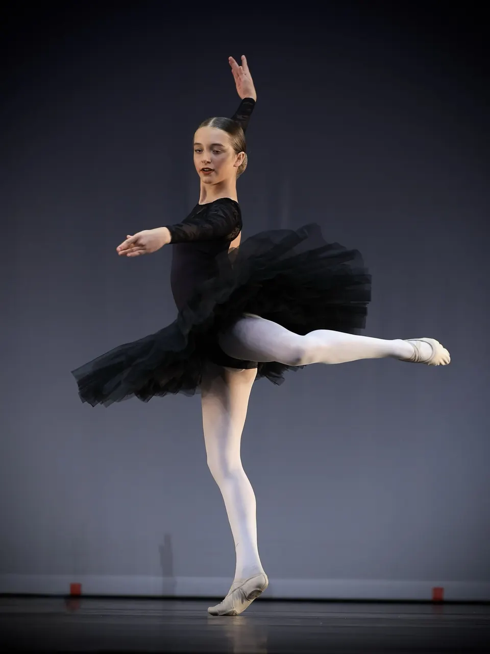 Manon Maindivide dance performance in a black tutu, balancing in arabesque on a dark stage