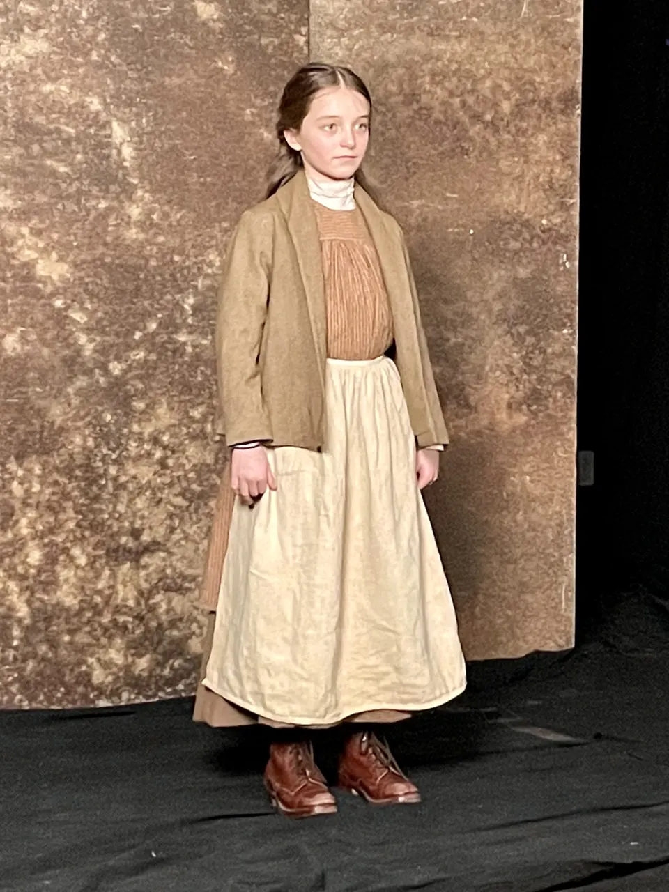 Manon Maindivide in period costume with a tan jacket and long cream skirt on a dark stage