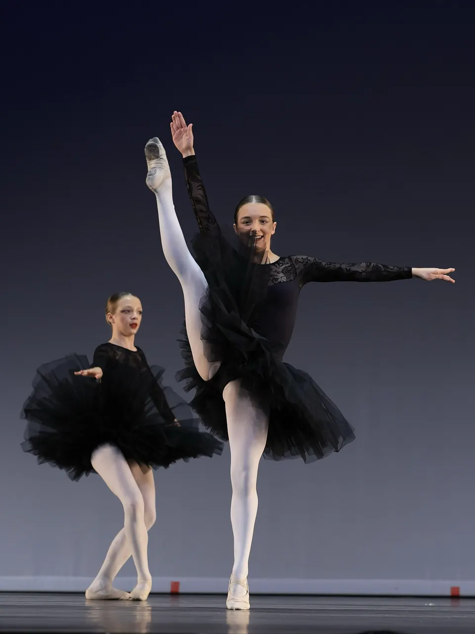 Manon Maindivide dance performance in a black tutu and white tights, high kick beside another ballerina