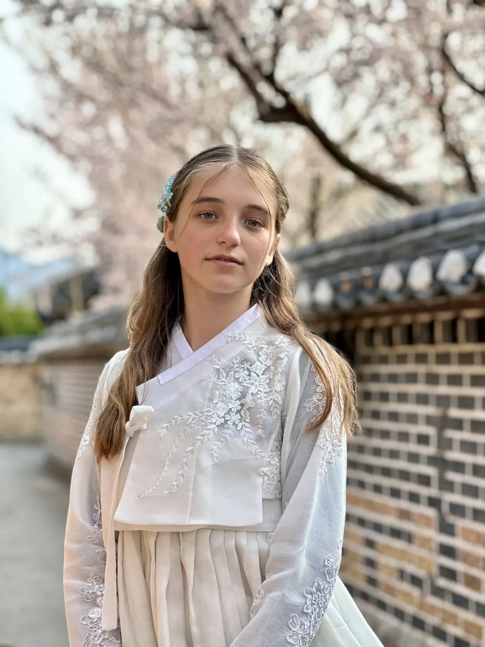 Manon Maindivide photo shoot in a white traditional jacket by a brick wall under spring blossoms