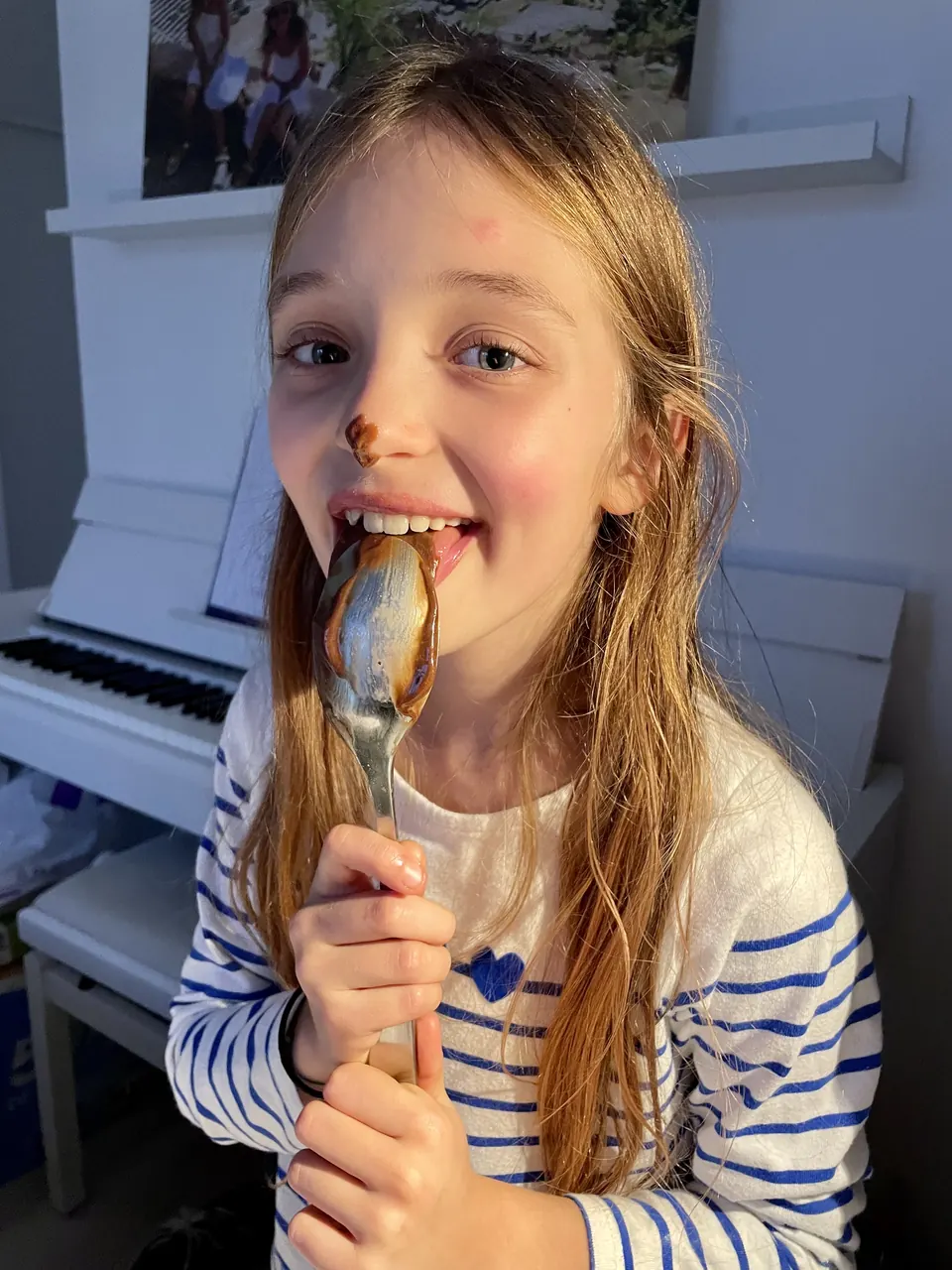 Manon Maindivide behind the scenes in a striped top, playfully biting a spoon near a white piano