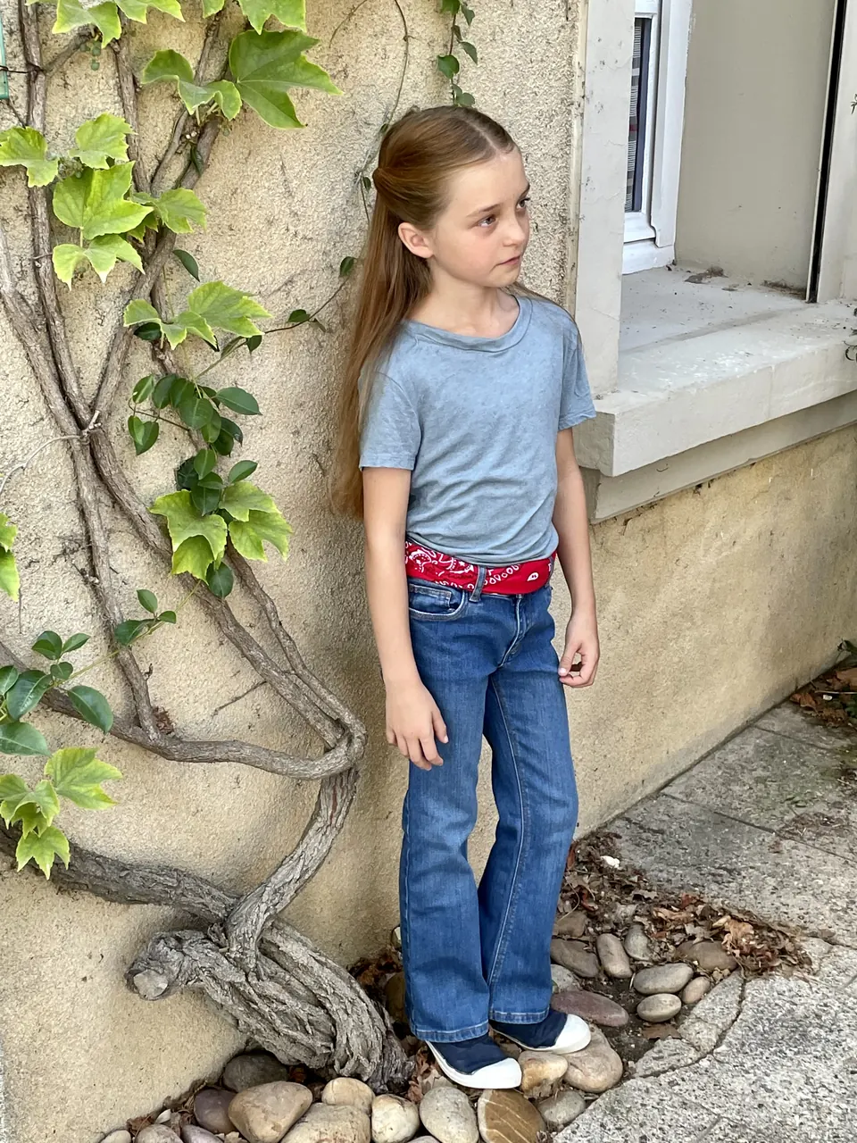 Manon Maindivide photo shoot in a blue tee and flared jeans, standing by an ivy-covered wall
