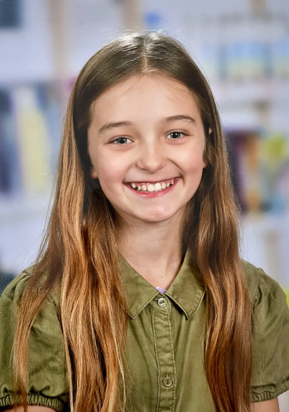 Manon Maindivide headshot with a wide smile, wearing an olive green shirt in front of a blurred bookshelf