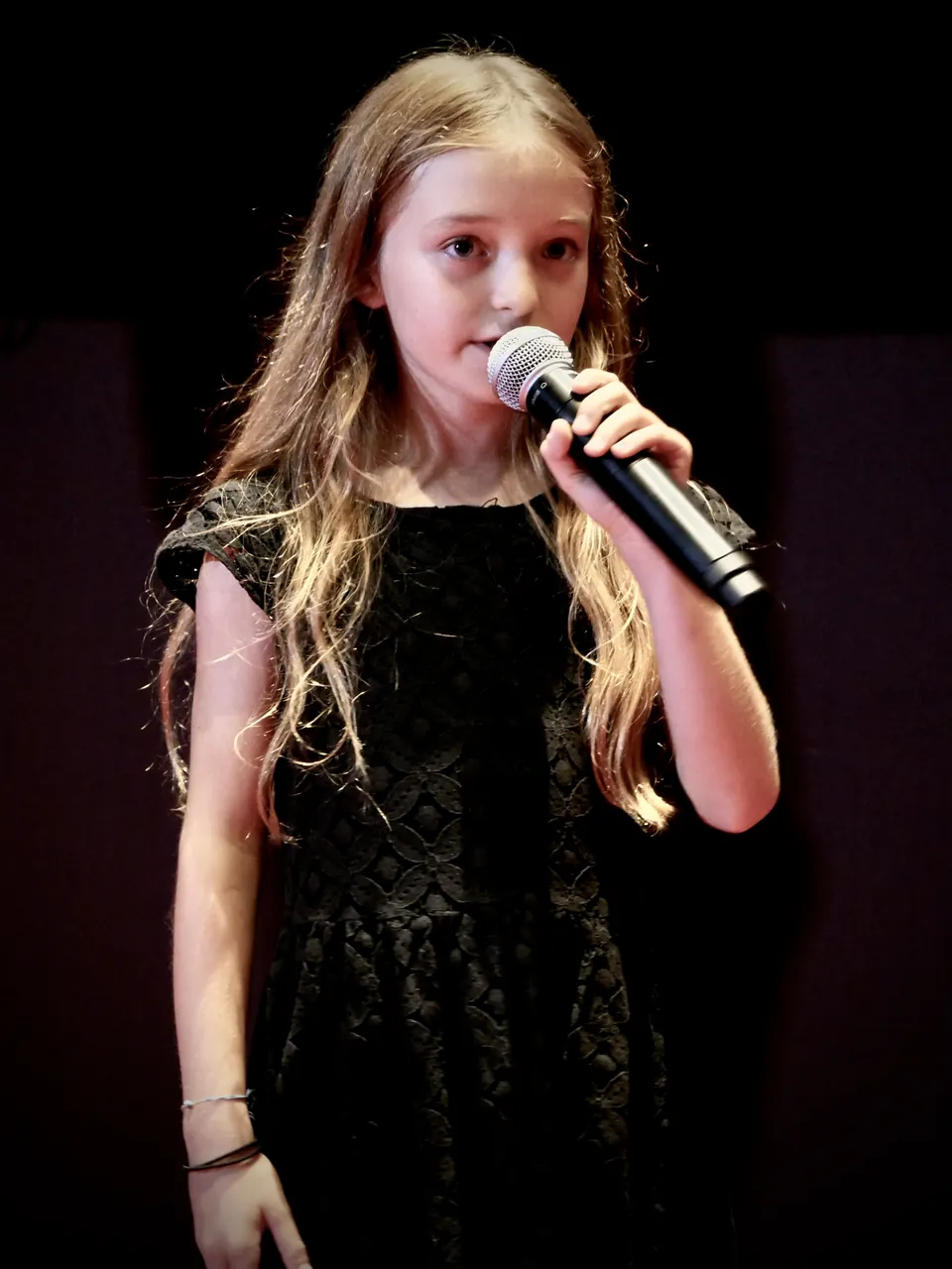 Manon Maindivide on stage singing into a microphone, wearing a black dress against a dark backdrop