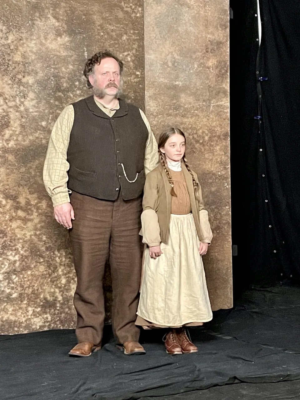Manon Maindivide behind the scenes in a period costume, standing beside an actor in a brown waistcoat