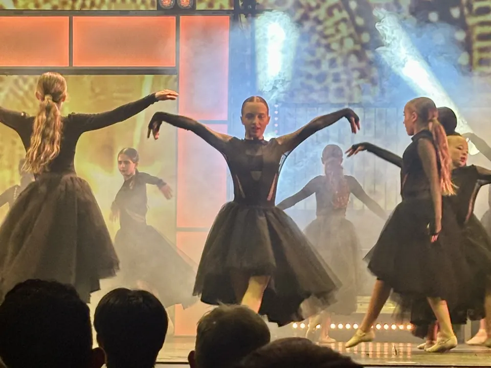 Manon Maindivide dance performance in a black tutu, arms spread wide with other dancers under stage lights