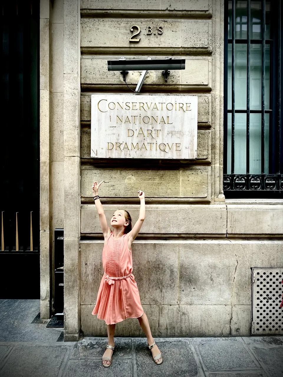 Manon Maindivide casual photo in a coral dress, arms raised in front of a historic stone building