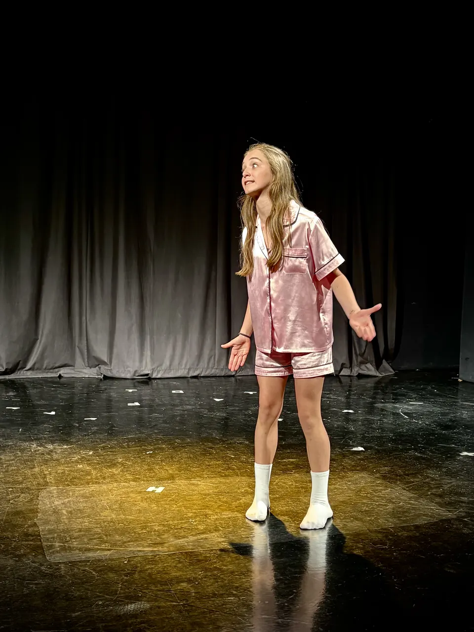 Manon Maindivide on stage in pink satin pyjamas and white socks, arms open during a theatre scene