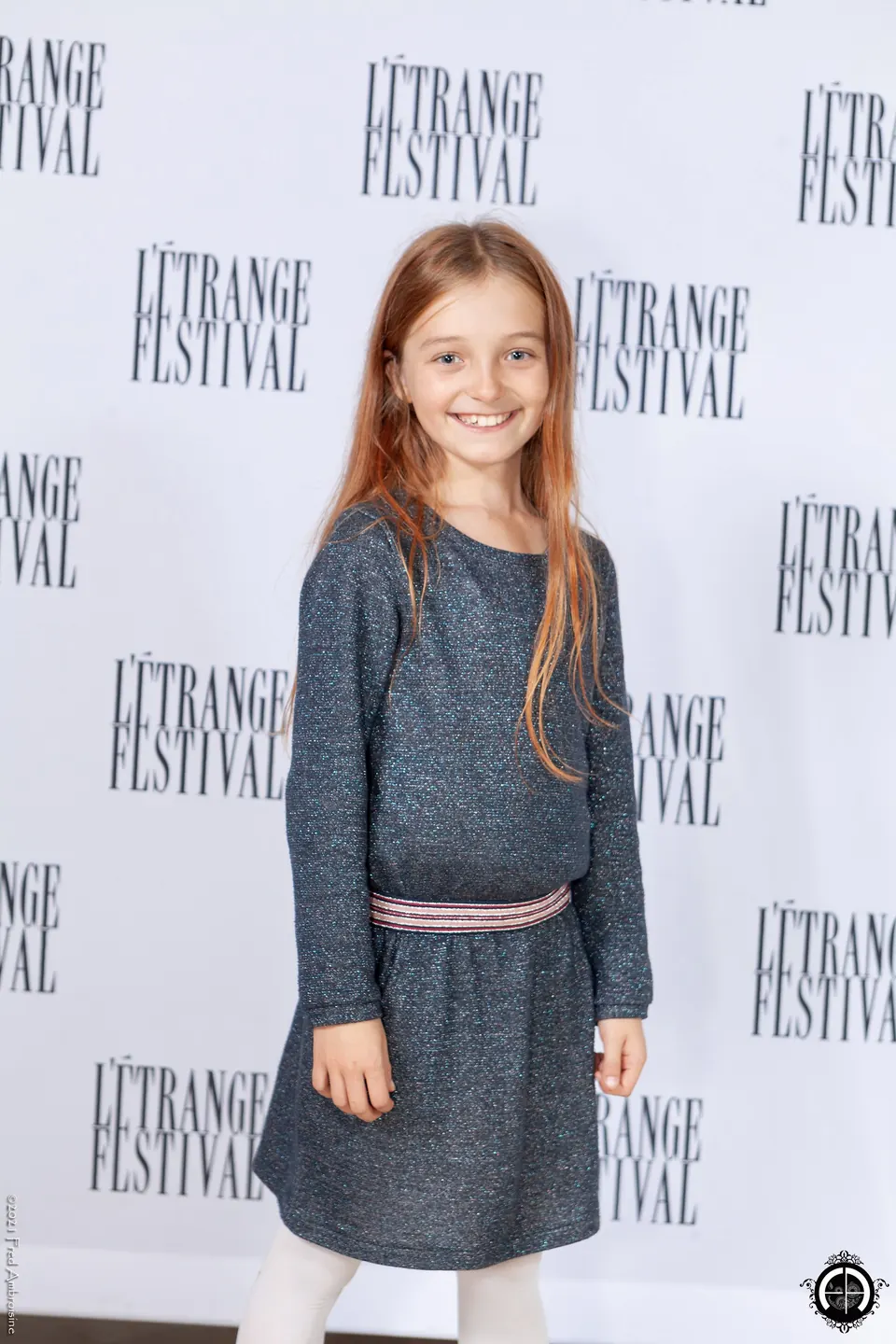 Manon Maindivide at a festival photo wall, smiling in a grey knit dress with a striped belt
