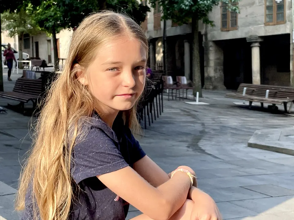 Manon Maindivide casual photo crouching in a sunny paved square, long hair over her shoulder