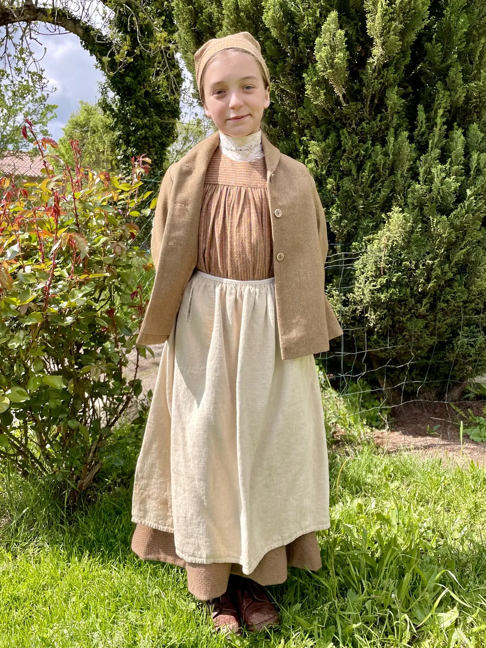 Manon Maindivide in period costume with a headscarf, apron and jacket in a garden