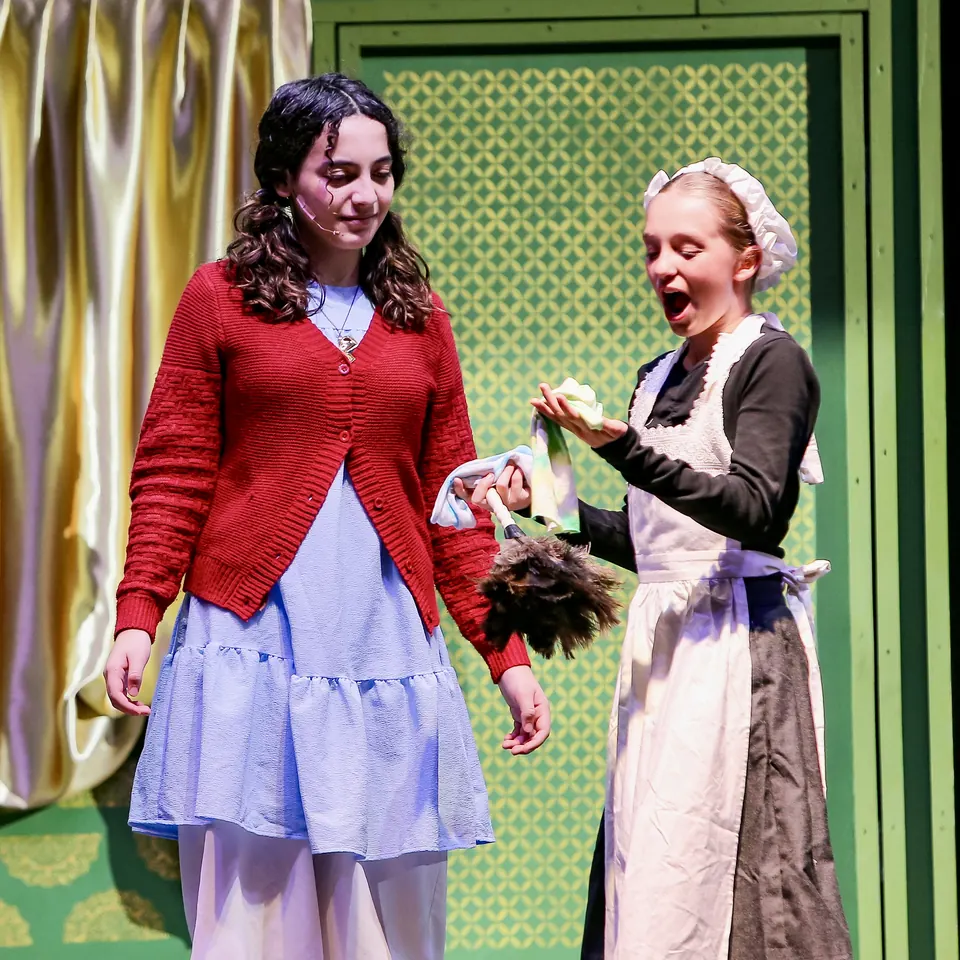 Manon Maindivide on stage in a red cardigan and blue dress beside an actress in a maid costume