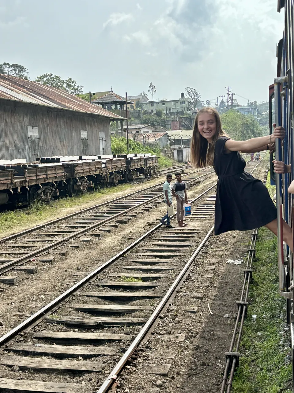 Manon Maindivide on set in a black dress, leaning out from a train beside old railway tracks
